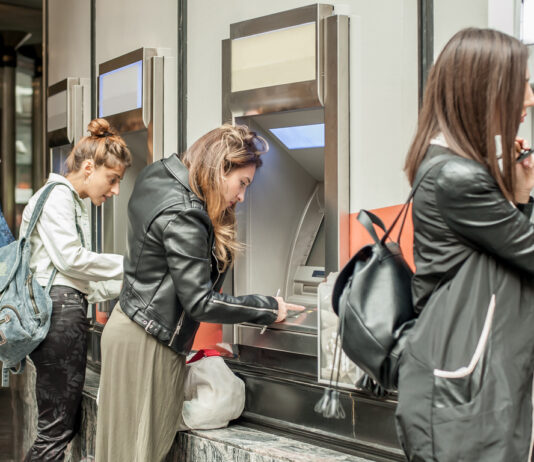 Frauen an einem Geldautomaten in einer Bank. Sie stehen jede an einem eigenen Automaten, tippen ihren Pin ein oder warten auf die Geldbewegung oder Auszahlung. Die Filiale ist hell beleuchtet.