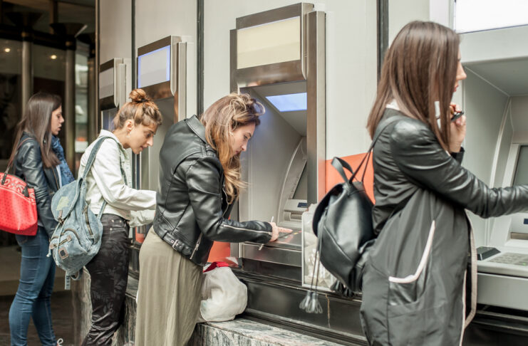 Frauen an einem Geldautomaten in einer Bank. Sie stehen jede an einem eigenen Automaten, tippen ihren Pin ein oder warten auf die Geldbewegung oder Auszahlung. Die Filiale ist hell beleuchtet.