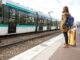 Eine junge Frau wartet am Bahnsteig mit einem Koffer auf die Deutsche Bahn