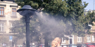 Ein Mann kühlt sich von der sommerlichen Hitze ab. Er steht im Park unter einer Düse, die leichten Sprühregen von sich gibt. Im Hintergrund sitzt ein Mann auf einer Parkbank unter einem Baum. Es ist ein heißer Sommertag.