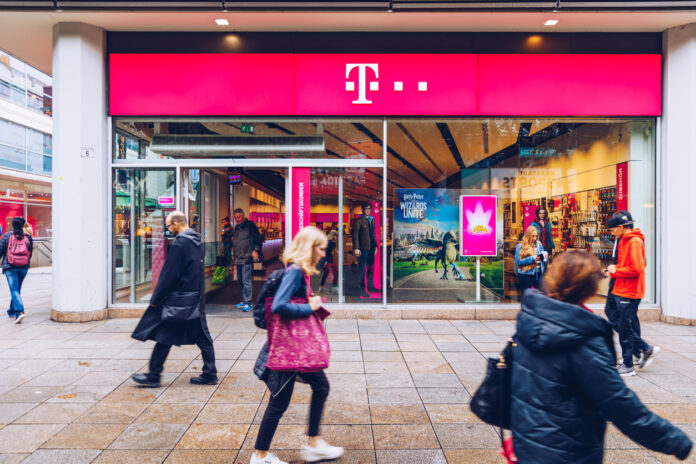 Eine Telekom-Filiale in einer Innenstadt. Der Laden des Handy- und Telefonanbieters ist in dem klassisch pinken Telekom-Farbton gehalten. An dem Geschäft laufen mehrere Passanten vorbei.