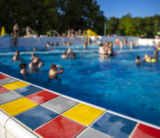 Verschiedene Badegäste schwimmen im Freibad. Die Sonne scheint und das Wasser ist blau. Um den Pool herum sind bunte Fliesen.