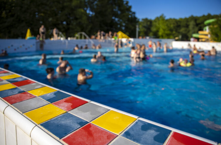 Verschiedene Badegäste schwimmen im Freibad. Die Sonne scheint und das Wasser ist blau. Um den Pool herum sind bunte Fliesen.