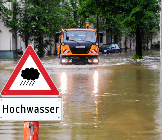 Ein Lkw fährt über eine überschwemmte Straße. Das Scheinwerferlicht ist an und die Räder des Lkws kämpfen sich durch die Wassermassen. Im Vordergrund ist ein Warnschild vor Hochwasser zu erkennen.