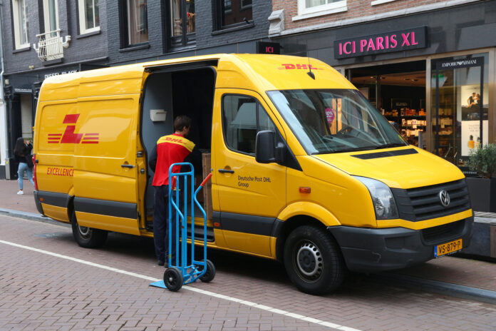 Ein DHL-Bote steht an seinem gelben Fahrzeug. Er holt gerade Post oder Pakete aus dem Transporter und will sie zustellen. Daneben steht ein blauer Rollwagen.