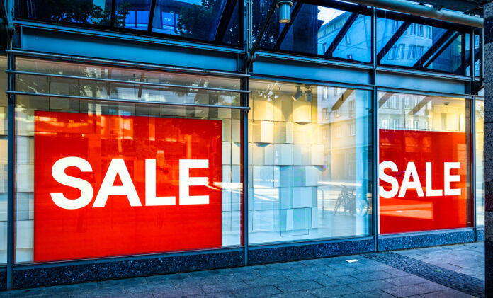 An einem Geschäft in der Innenstadt sind im Schaufenster große rot-weiße Schilder mit der Aufschrift SALE angebracht. Diese Werbung soll die Kunden auf Angebote eines Ausverkaufs hinweisen und in den Laden locken.