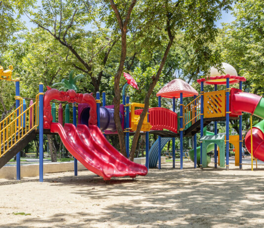 Bunter Spielplatz im Park bei Sonnenschein. Der Spielplatz besteht aus Klettergerüsten, einer Rutsche und einer Schaukel. Die Spielgeräte sind aus leichtem und robustem Kunststoff. Der Spielplatz ist für kleine und große Kinder geeignet.