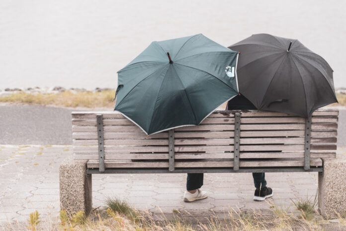 Zwei Personen sitzen mit Regenschirmen auf einer Bank