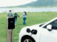 im Vordergrund lädt ein Elektroauto und im Hintergrund laufen Menschen auf einen See zu
