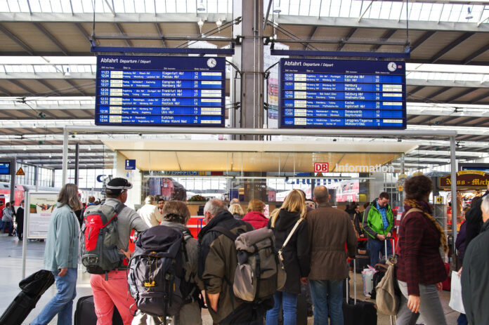Reisende warten am Bahnhof und schauen die Verbindungstafel an