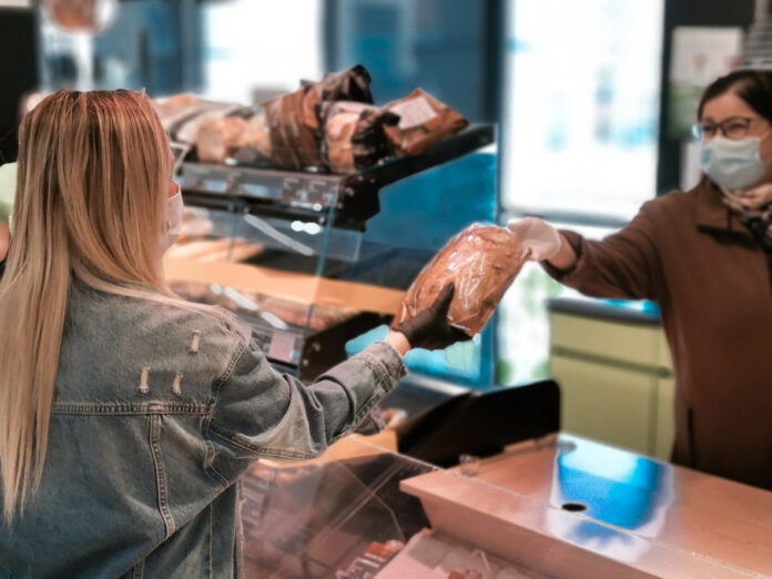 Blonde Frau steht an der Theke und kauft ein Brot in einer Tüte. Die Verkäuferin und Kundin tragen beide Handschuhe.