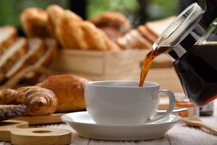 Aus einer Kaffeekanne wird das Getränk in eine weiße Tasse mit Untertasse eingeschänkt