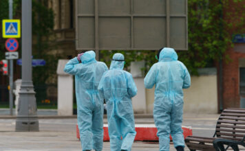 Vier Personen in blauen Schutzanzügen gehen durch eine menschenleere Straße. Sie reinigen die öffentlichen Plätze von Viren und Bakterien und desinfizieren alles.