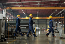 Zwei Männer und eine Frau, offensichtlich angestellte Mitarbeiter, laufen in blauer Schutzkleidung und mit gelben Helmen durch eine große Produktionshalle.