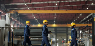 Zwei Männer und eine Frau, offensichtlich angestellte Mitarbeiter, laufen in blauer Schutzkleidung und mit gelben Helmen durch eine große Produktionshalle.