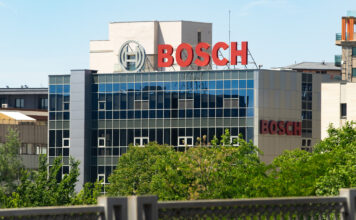 Das Bosch Gebäude hinter einer Begrünung mit Bäumen und einer Abgrenzung. Die Glasfronten scheinen im Sonnenlicht und reflektieren ein gegenüberliegendes Gebäude. Auf dem Dach ist das rote Bosch Logo klar sichtbar.