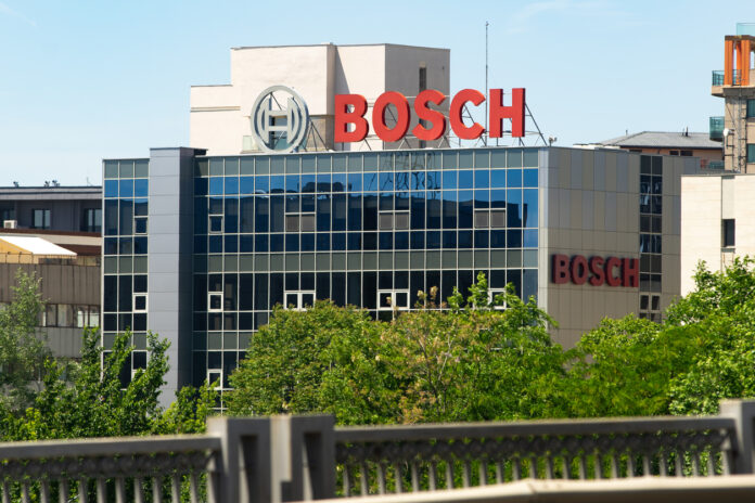 Das Bosch-Gebäude hinter einer Begrünung mit Bäumen und einer Abgrenzung. Die Glasfronten scheinen im Sonnenlicht und reflektieren ein gegenüberliegendes Gebäude. Auf dem Dach ist das rote Bosch-Logo klar sichtbar.