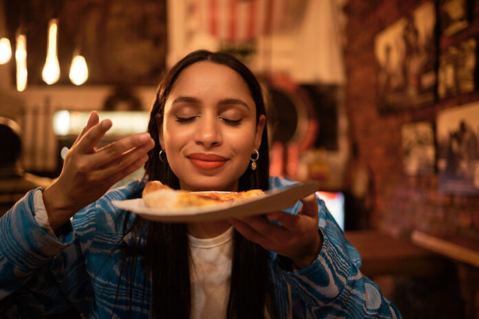 Eine Frau riecht an einem Stück Pizza vor einem Imbiss