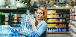 Eine Frau mit einem besorgten Gesichtsausdruck hebt ein Wasserpaket in einem Supermarkt hoch.