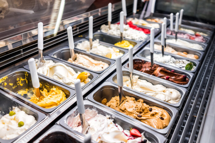 Die Auslage mit Speiseeis in einem Eiscafe. Die verschiedenen Eissorten stehen bereit für den Außer-Haus-Verkauf. Die Kunden stehen an und bekommen dann eine Kugel Eis aus dem Eiscafe in der Waffel.