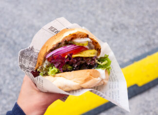 An einem hiesigen Foodtruck bekommen Kunden leckere hausgemachte Burger, Döner und anderes Fast Food mit Salat, Fleisch, Fleischersatz, Gemüse und Soße. Ein Kunde verspeist seinen Imbiss auf der Straße.