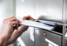 Ein Briefkasten aus Metall, für den man einen Schlüssel benötigt ist zu sehen. Eine Hand öffnet die Klappe des Kastens und schiebt weiße Briefe in den Briefkasten hinein.