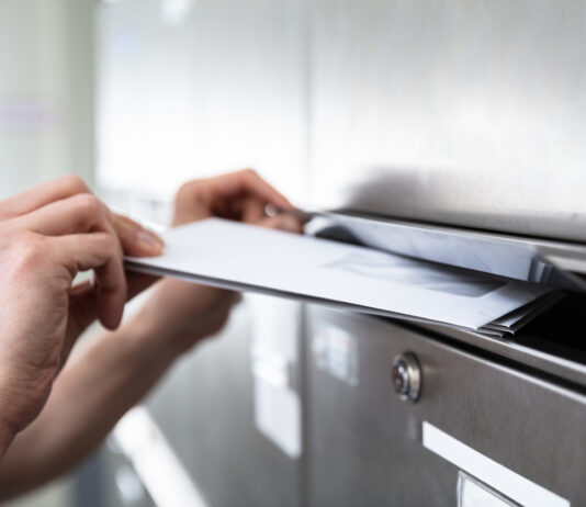 Ein Briefkasten aus Metall, für den man einen Schlüssel benötigt ist zu sehen. Eine Hand öffnet die Klappe des Kastens und schiebt weiße Briefe in den Briefkasten hinein.