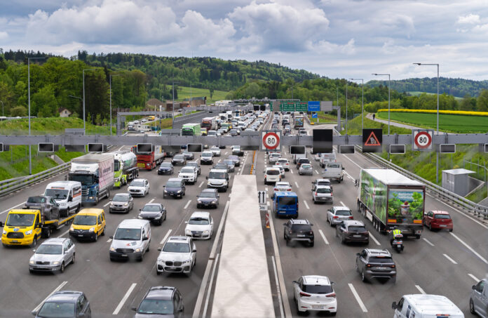 Zwei jeweils vierspurige Autobahnen in Deutschland, die in entgegengesetzte Richtungen dicht befahren sind. Die Autos und Lkw fahren im Stop-and-Go, weil ein Stau bevorsteht. Über der Fahrbahn ist ein Tempolimit und ein Baustellen-Schild angebracht. Im Hintergrund ist schöne grüne Landschaft.