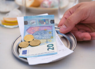 Ein Gast reicht mit der rechten Hand auf einem kleinen Silbertablett den Rechnungsbetrag hin. Der Kassenzettel liegt unter einem 20-Euro-Schein und einigen Münzen. Im Hintergrund sieht man noch Gläser und Teller.