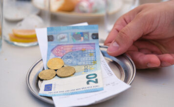 Ein Gast reicht mit der rechten Hand auf einem kleinen Silbertablett den Rechnungsbetrag hin. Der Kassenzettel liegt unter einem 20-Euro-Schein und einigen Münzen. Im Hintergrund sieht man noch Gläser und Teller.