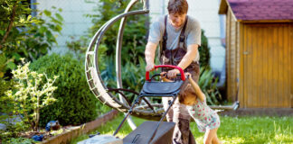 Ein Vater mäht mit seiner kleinen Tochter den Rasen im Garten. Das kleine Mädchen versucht den Rasenmäher anzuschieben, während der Vater ihn festhält.