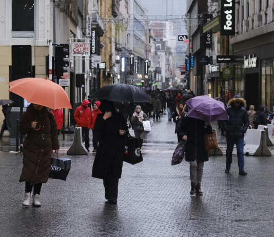 Menschen mit Regenschirmen auf einer verregneten Straße in einer größeren Stadt. Trotz des schlechten Wetters ist es sehr belebt. Der Himmel am Horizont ist grau.