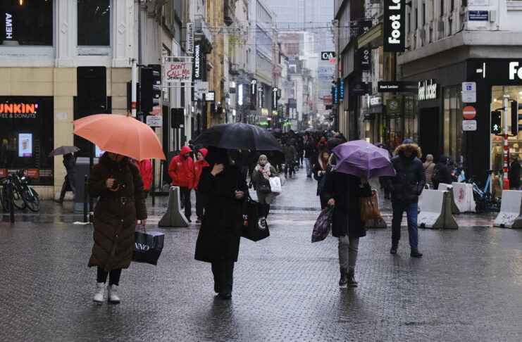 Menschen mit Regenschirmen auf einer verregneten Straße in einer größeren Stadt. Trotz des schlechten Wetters ist es sehr belebt. Der Himmel am Horizont ist grau.