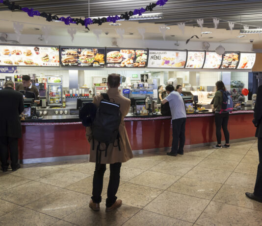 Mehrere Gäste stehen bei McDonald's an der Kasse an, um Fastfood aus dem Angebot auf den Leuchtanzeigen zu bestellen.
