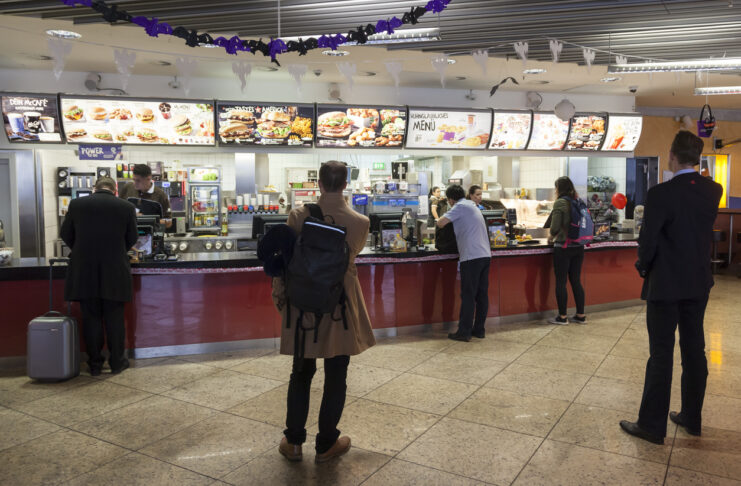 Mehrere Gäste stehen bei McDonald's an der Kasse an, um Fastfood aus dem Angebot auf den Leuchtanzeigen zu bestellen.