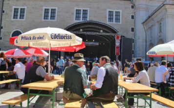 Mehrere Bierbänke stehen außen in der Sonne vor einer Bühne eines gut bürgerlichen Straßenfestes. In der Mitte sind zwei Männer in Tracht gekleidet. Nebenan sitzen weitere Gäste unter Sonnenschirmen mit Getränken.
