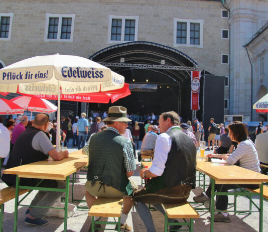 Mehrere Bierbänke stehen außen in der Sonne vor einer Bühne eines gut bürgerlichen Straßenfestes. In der Mitte sind zwei Männer in Tracht gekleidet. Nebenan sitzen weitere Gäste unter Sonnenschirmen mit Getränken.