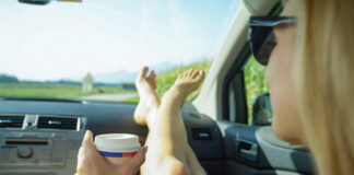 Ein junge blonde Frau mit Sonnenbrille sitzt auf dem Beifahrersitz eines Autos während eines Road-Trips und streckt ihre Füße mit einem Kaffeebecher in der Hand auf dem Armaturenbrett aus.