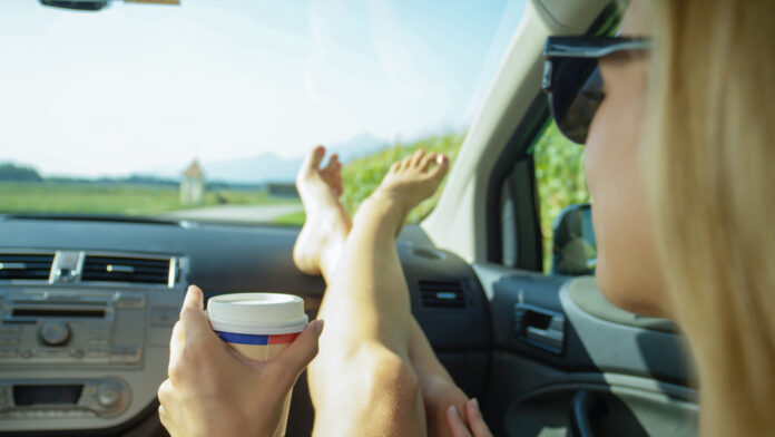 Ein junge blonde Frau mit Sonnenbrille sitzt auf dem Beifahrersitz eines Autos während eines Road-Trips und streckt ihre Füße mit einem Kaffeebecher in der Hand auf dem Armaturenbrett aus.