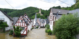 Ein Dorf mit alten Fachwerkhäusern ist überflutet. Die Straßen sind voller Wasser und der Wasserpegel ist so hoch, dass sie nicht mehr begehbar sind. Das Dorf befindet sich in einem Tal; im Hintergrund sieht man einige Hügel.