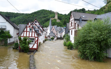 Ein Dorf mit alten Fachwerkhäusern ist überflutet. Die Straßen sind voller Wasser und der Wasserpegel ist so hoch, dass sie nicht mehr begehbar sind. Das Dorf befindet sich in einem Tal; im Hintergrund sieht man einige Hügel.