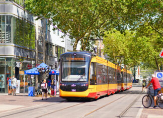 Eine viel befahrene Straße in einer Innenstadt mit Bahn und Passanten. Grüne Bäume pflastern die Allee, an der sich Geschäfte und Cafés anreihen, Menschen vorbeigehen und die Straßenbahn vorbeifährt.