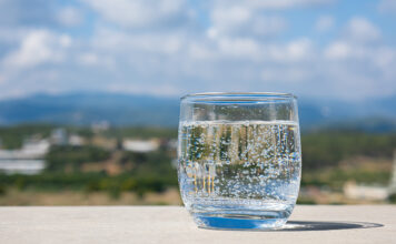 Ein Wasserglas mit Mineralwasser. Es steht auf einem Tisch aus Beton und im Hintergrund zeigt sich eine malerische Landschaft mit blauem Himmel. Die Kohlensäure perlt im Wasser.