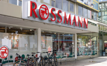 Der Eingangsbereich einer Rossmann-Filiale. Über der Eingangstür steht der Name der Drogerie und das Logo. Die Glastür öffnet sich automatisch und gibt den Blick auf das Sortiment und den Kassenbereich frei.
