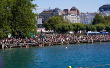 Junge Leute feiern am Wasser. Im Vordergrund ist ein großer See, dahinter eine Menschenmenge auf Treppen. Die Menschen sitzen vor einem großen Gebäude.
