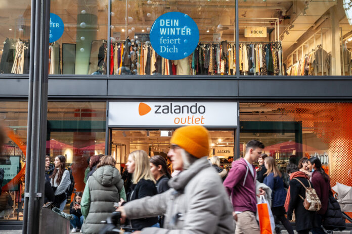 Selektive Unschärfe auf einem Zalando-Logo auf ihrem Outlet-Store in Köln mit verschwommenen Kunden, die vorbeigehen.