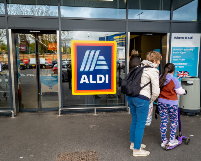 Eine Frau und ein Kind stehen vor dem Eingangsbereich einer Aldi-Süd-Filiale. Die Frau packt etwas in den Rucksack des Kindes. Sie hat selbst einen Rucksack auf dem Rücken.