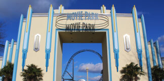 Der Eingang zum Movie Park erstrahlt an einem sommerlichen, warmen und sonnigen Tag unter blauem Himmel. Im Hintergrund ist eine berühmte Attraktion zu sehen.