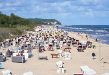 Urlauber am überfüllten Strand an der deutschen Ostseeküste. An dem weiten Strand liegen viele Menschen und sonnen sich. Im Hintergrund sieht man grüne Landschaft und das Meer.