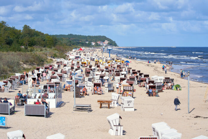 Urlauber am überfüllten Strand an der deutschen Ostseeküste. An dem weiten Strand liegen viele Menschen und sonnen sich. Im Hintergrund sieht man grüne Landschaft und das Meer.